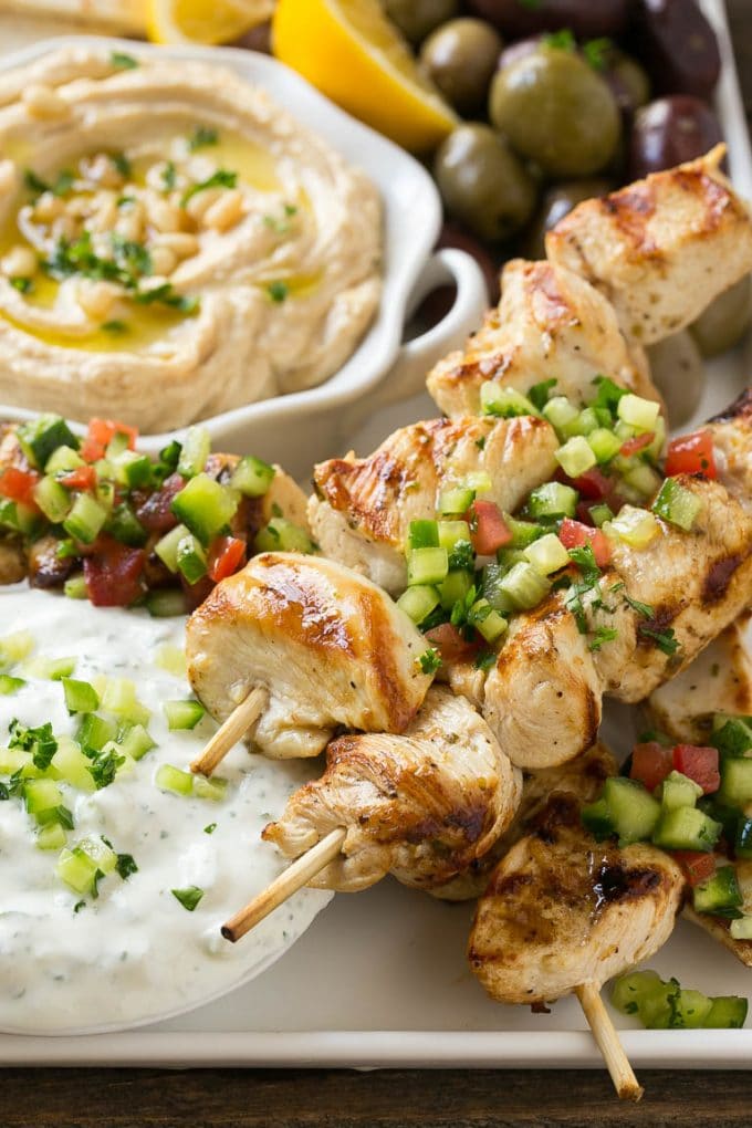 Greek Chicken Souvlaki Recipe | Easy Greek Chicken | Chicken Souvlaki | Greek Chicken Skewers | Easy Greek Chicken Skewers #chicken #kabobs #glutenfree #keto #lowcarb #grilling #fuyos A tray of chicken souvlaki kabobs with olives, hummus, pita bread and yogurt sauce.