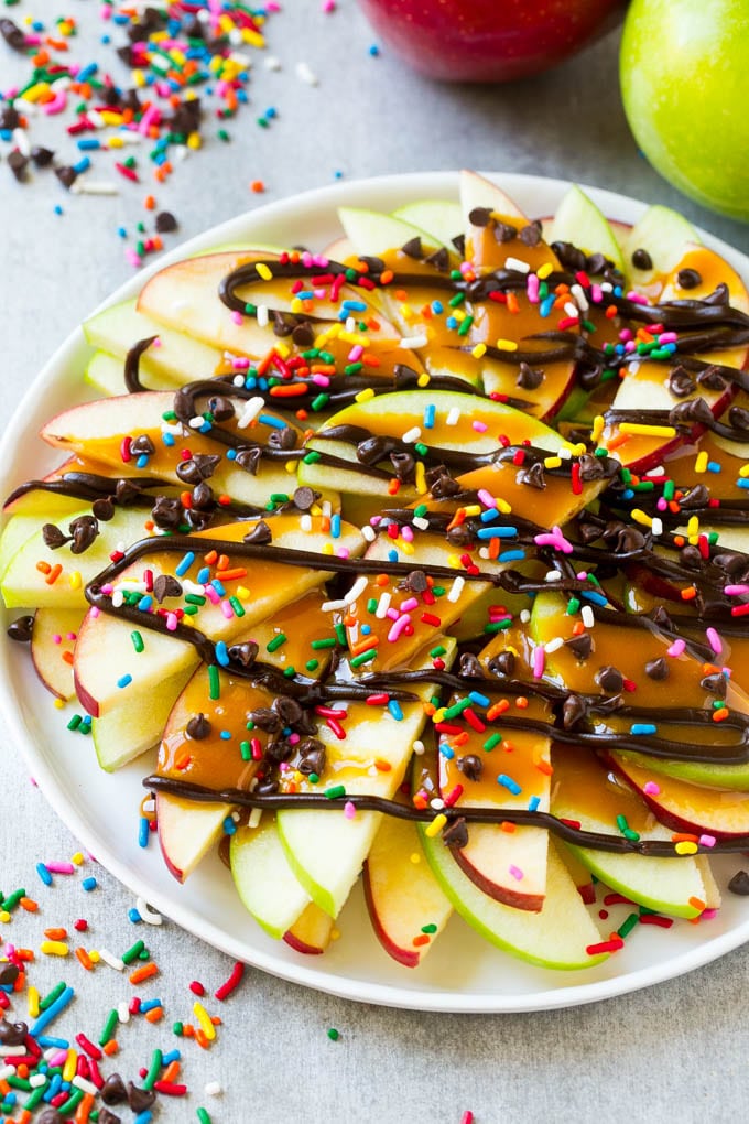 Apple Nachos Recipe | After School Snack | Caramel Apples #apple #apples #caramelapple #snack #dessert #fuyos A plate of sliced apple nachos topped with caramel and chocolate sauces, rainbow sprinkles and mini chocolate chips