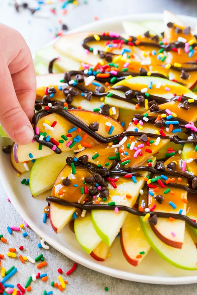 Apple Nachos Recipe | After School Snack | Caramel Apples #apple #apples #caramelapple #snack #dessert #fuyos A plate of caramel apple nachos with a hand reaching out to take one.