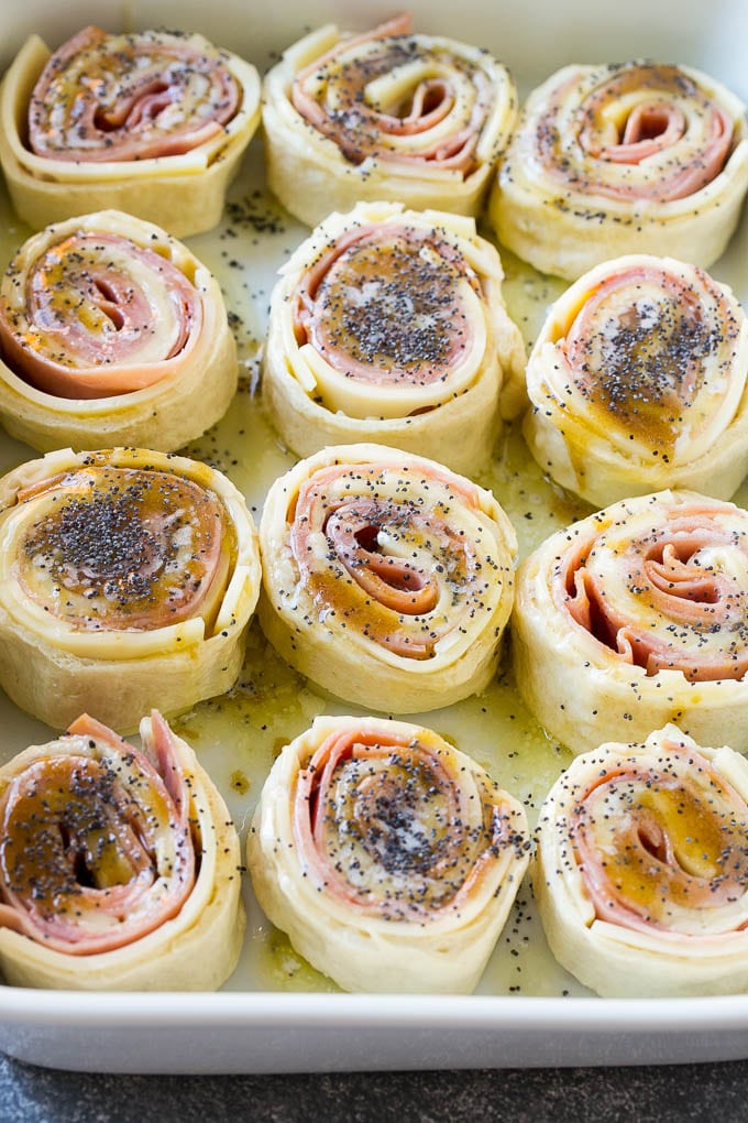 Ham and Cheese Pinwheels Recipe | Ham and Cheese Roll Ups | Ham and Cheese Rolls | Ham and Cheese Bread Rolls #ham #cheese #appetizer #pinwheels #fuyos Dough rolled up with ham and cheese and topped with a poppy seed butter in a ceramic dish.