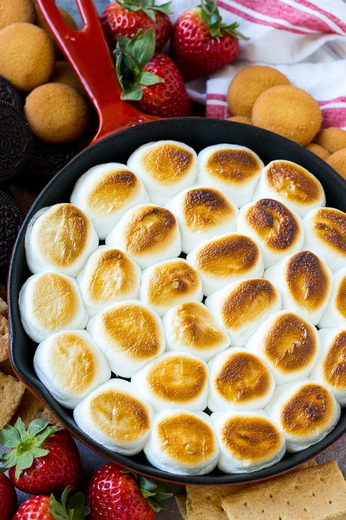 S'mores Dip Recipe | S'mores Skillet | Baked S'mores #smores #dip #chocolate #marshmallow #dessert #fuyos A skillet of s'mores dip with melted chocolate chips and toasted marshmallows, surrounded by graham crackers, cookies and strawberries.