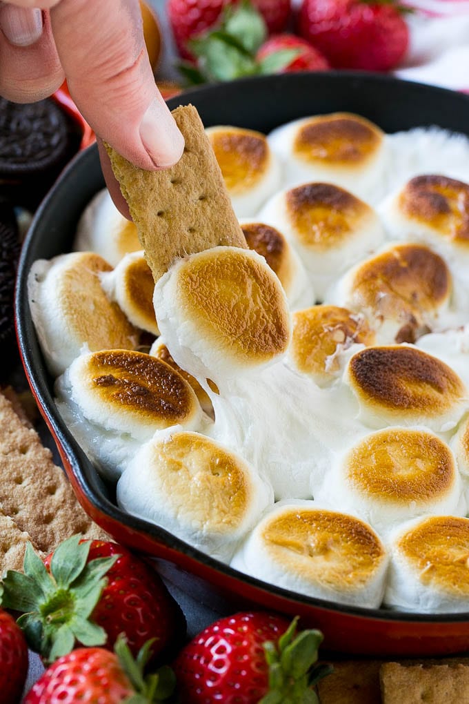 S'mores Dip Recipe | S'mores Skillet | Baked S'mores #smores #dip #chocolate #marshmallow #dessert #fuyos A graham cracker topped with melted marshmallow dipping into a skillet of s'mores dip.