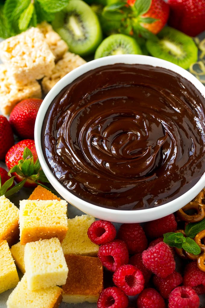 A bowl of chocolate fondue surrounded by fruit, cake, rice krispy treats and pretzels.