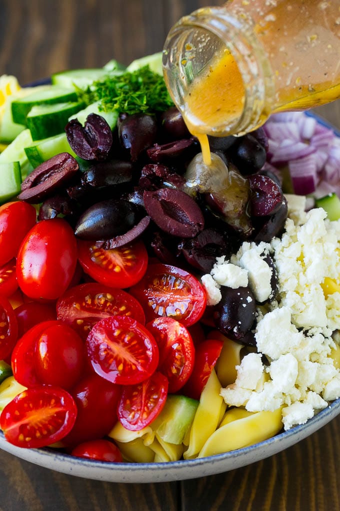 Greek Tortellini Salad Recipe | Pasta Salad | Tortellini Salad | Greek Salad #greek #tortellini #pasta #salad #cucumbers #olives #dinner #fuyos Salad dressing being poured over a bowl of tortellini and vegetables.