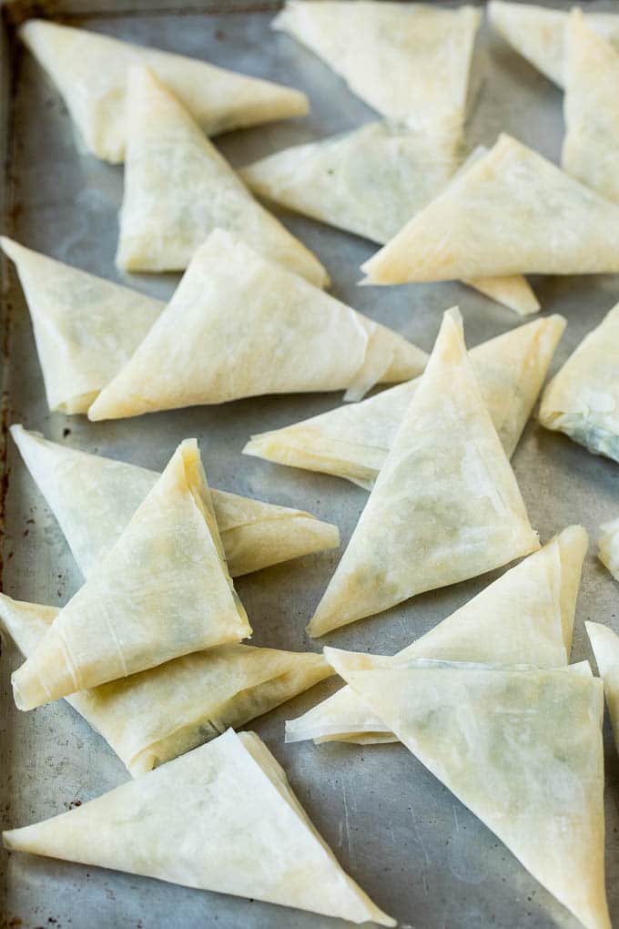 Spanakopita Recipe #appetizer #snack #spinach #cheese #fuyos Folded stuffed phyllo triangles on a sheet pan.