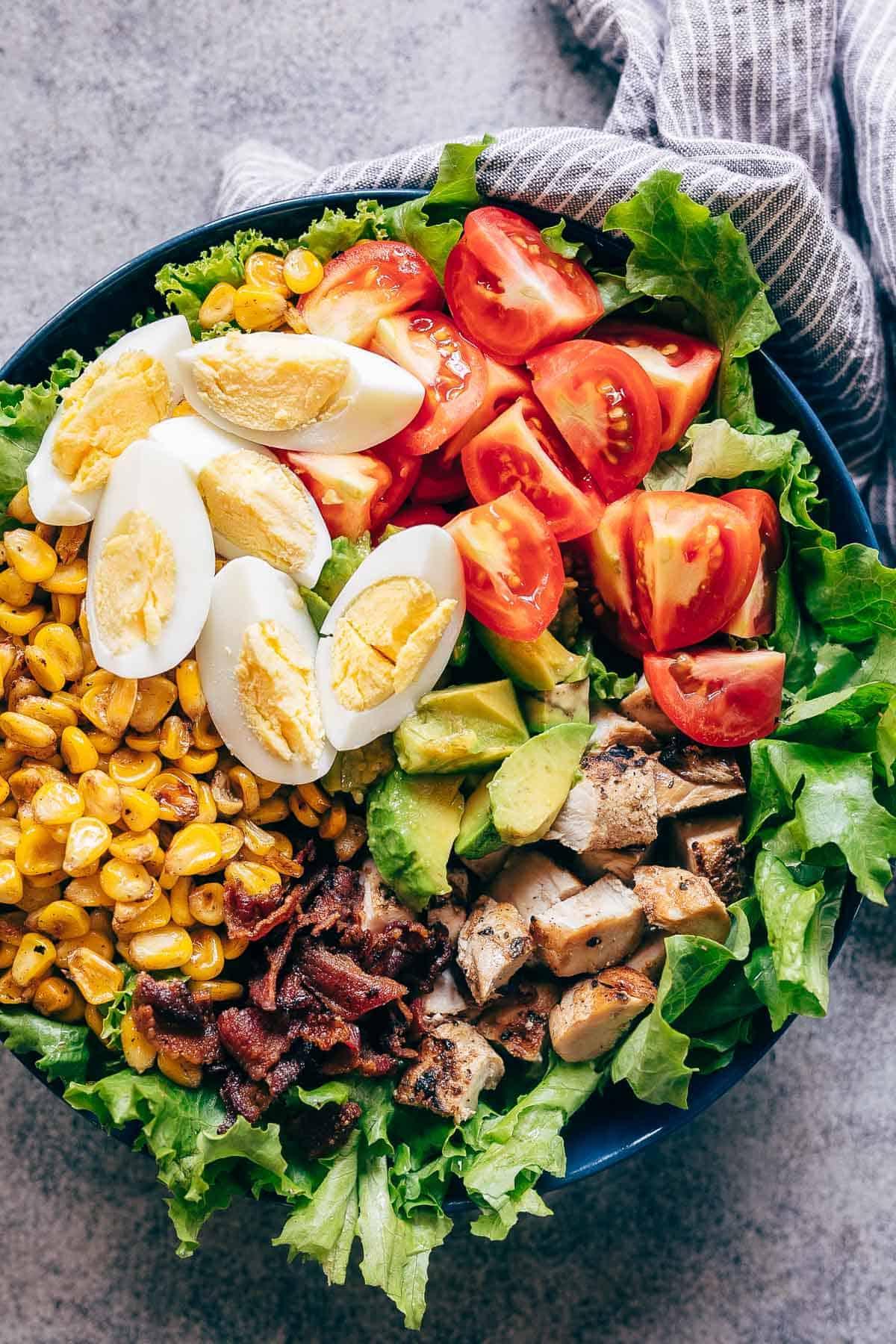 La classica ricetta dell'Insalata Cobb di Pollo, presentata con ingredienti freschi e disposti a strati: lattuga croccante, uova sode, mais grigliato, pancetta croccante, pollo grigliato, pomodori succosi e avocado cremoso. Servitela con una salsa vellutata al formaggio blu e avocado, a base di yogurt greco, per un pasto completo e delizioso. Perfetta per un pranzo sostanzioso o per una cena in compagnia. Prova anche varianti con bistecca o come piatto vegetariano. #insalata #ricetta #pollo #cena #facile #cobb #salutare Insalata Cobb di Pollo servita in una ciotola capiente, con lattuga fresca, pomodori maturi, pezzi di pollo grigliato, pancetta croccante, chicchi di mais dolce e uova sode, disposti a strati colorati e invitanti.