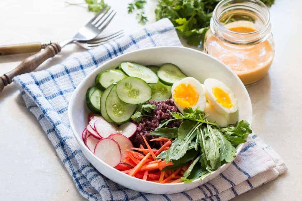 **Ciotola di Insalata di Riso Venere con Condimento Piccante alle Arachidi** 2 ciotola bianca con ingredienti per l'insalata di riso nero su una tovaglia blu e bianca, con forchette, coriandolo e un barattolo di salsa sul lato
