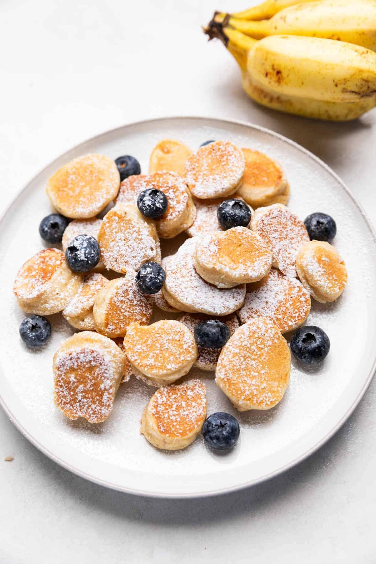 Mini Bocconcini di Pancake alla Banana 2 mini pancake di banana guarniti con zucchero a velo e mirtilli serviti su un piatto bianco
