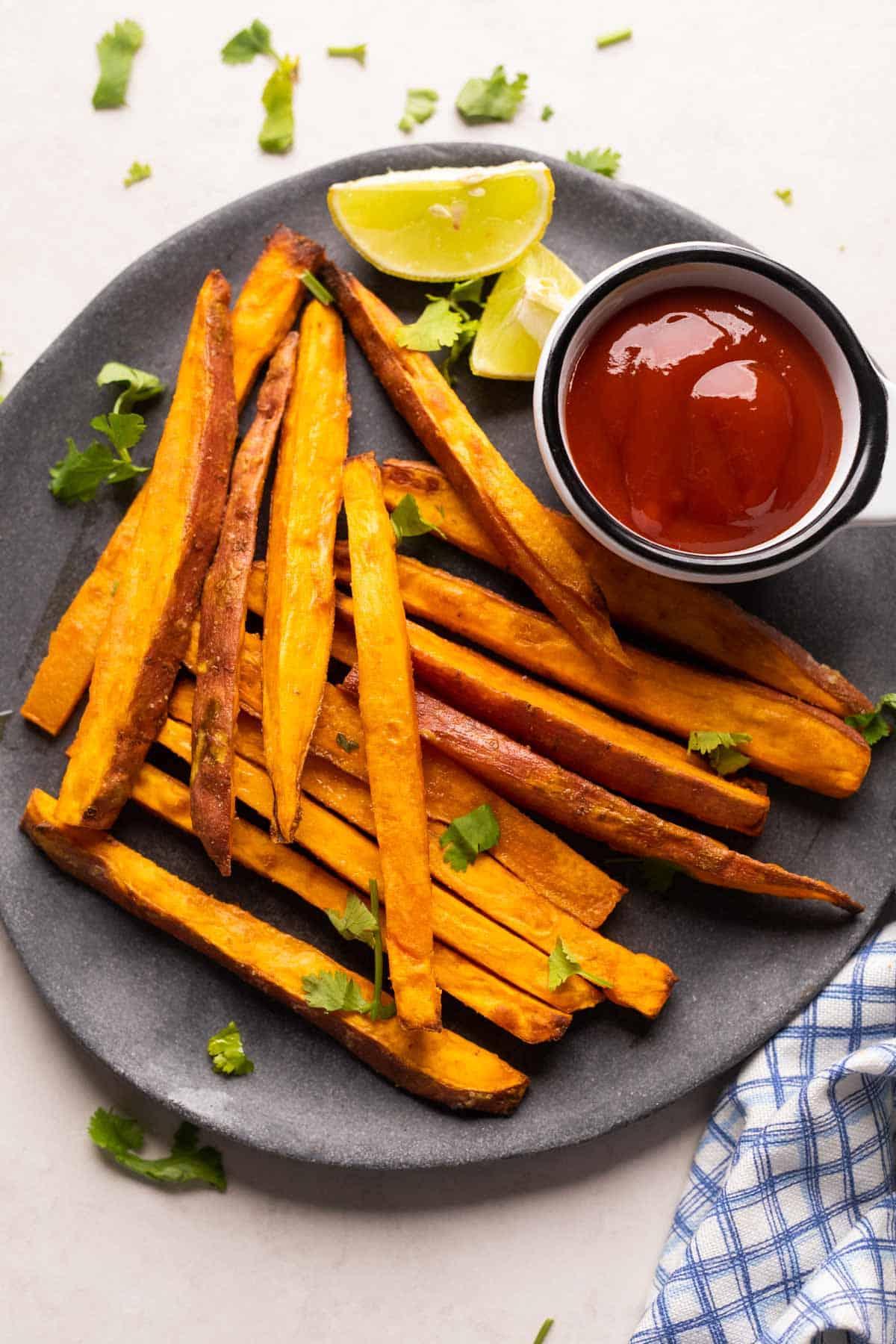 Patatine Dolci Croccantissime nella Friggitrice ad Aria 2 Immagine di patate dolci fritte cotte in friggitrice ad aria, servite su un piatto grigio con ketchup e spicchi di lime a lato