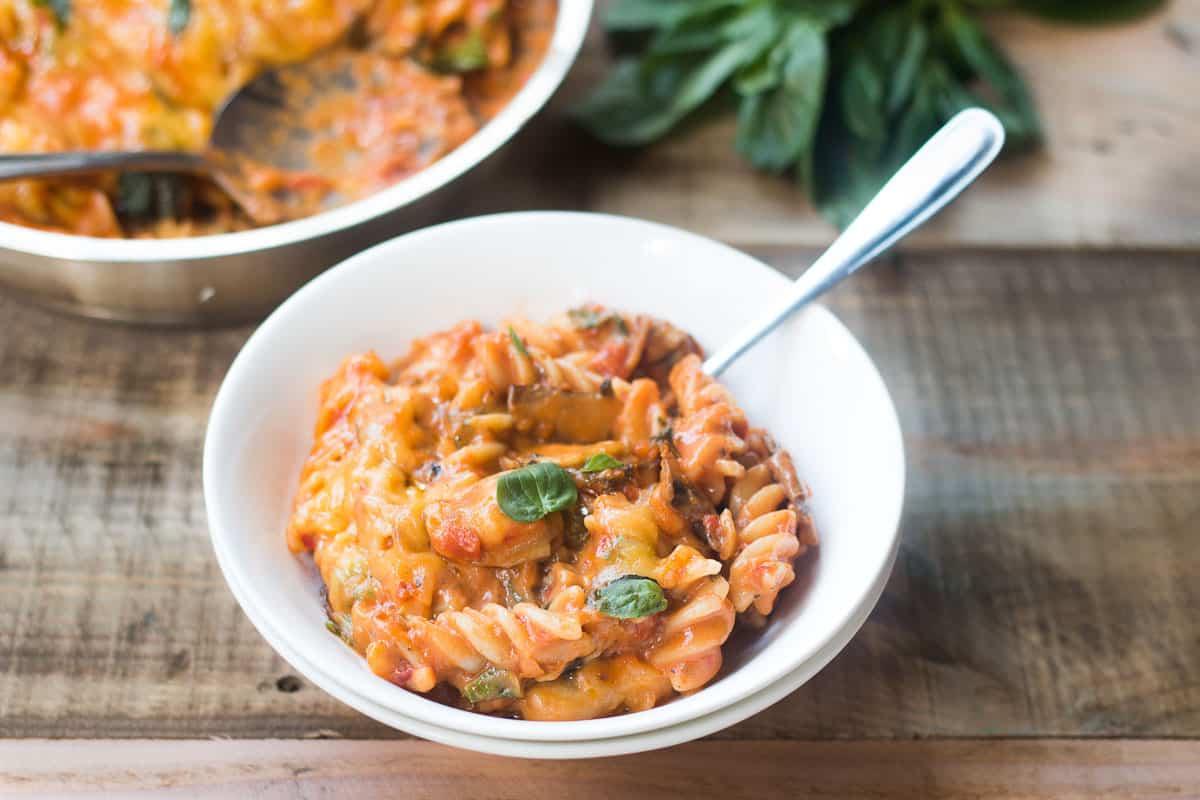 Pasta Domenicale di Verdure Tutto Incluso 2 Una confortante ciotola di pasta vegetariana della domenica con formaggio, guarnita con basilico fresco e servita in una ciotola bianca.