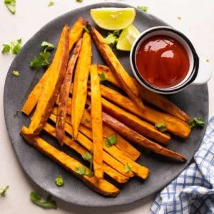 Patatine Dolci Croccantissime nella Friggitrice ad Aria 12 Immagine in miniatura di patate dolci fritte cotte in friggitrice ad aria, servite su un piatto grigio con ketchup e spicchi di lime a lato