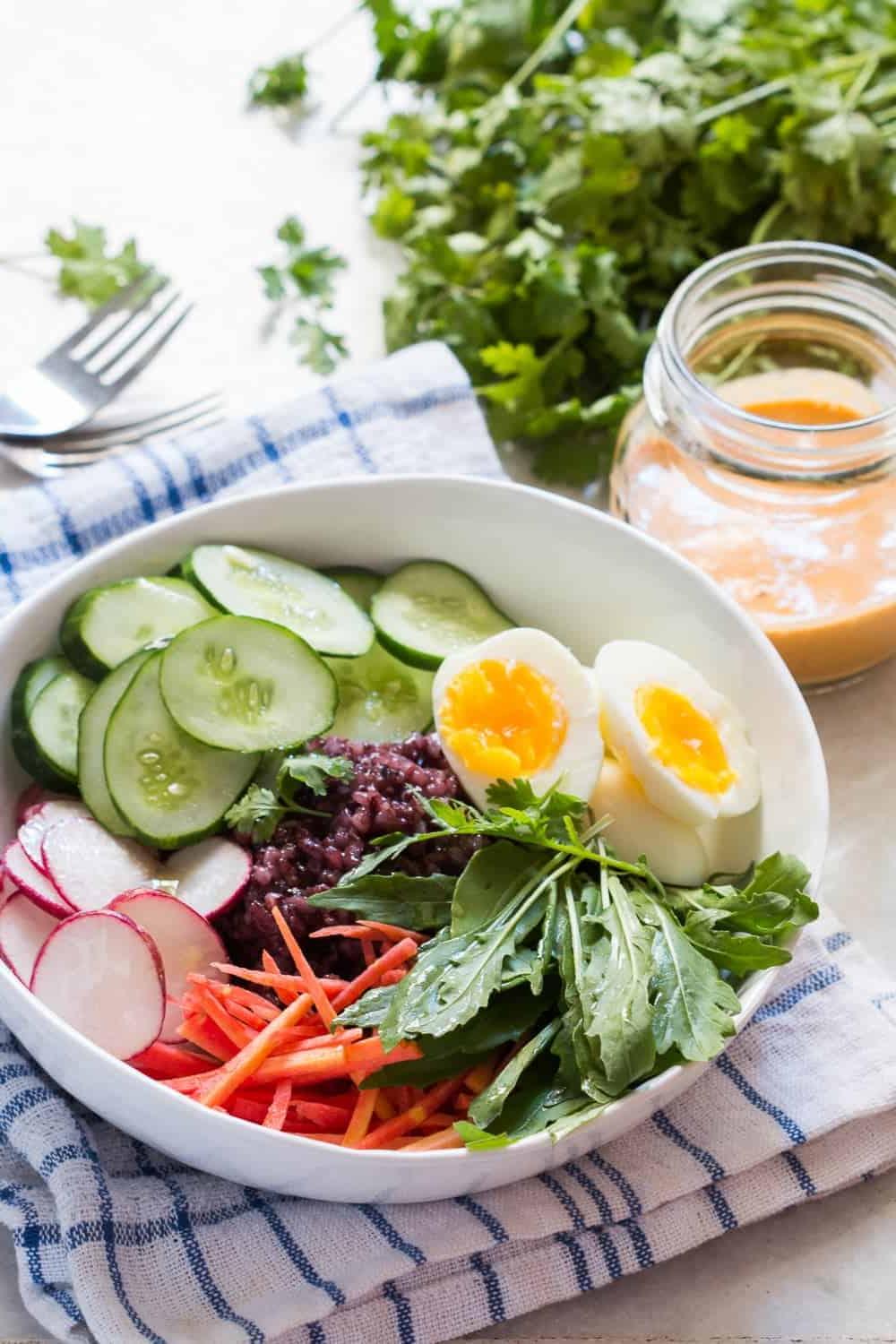 **Ciotola di Insalata di Riso Venere con Condimento Piccante alle Arachidi** 3 ciotola bianca con ingredienti per l'insalata di riso nero su una tovaglia blu e bianca, con forchette, coriandolo e un barattolo di salsa sul lato