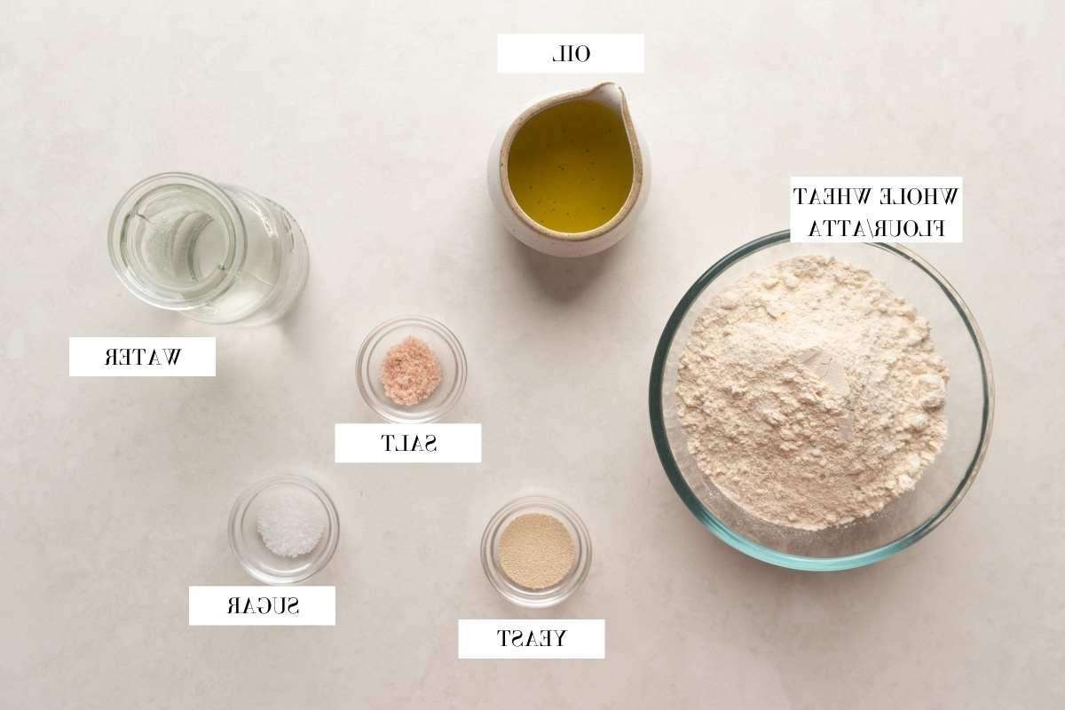 Ricetta del Pane Pita Integrale 3 Immagine ravvicinata di tutti gli ingredienti necessari per il pane pita integrale, chiaramente etichettati e disposti in modo ordinato su una superficie di legno.