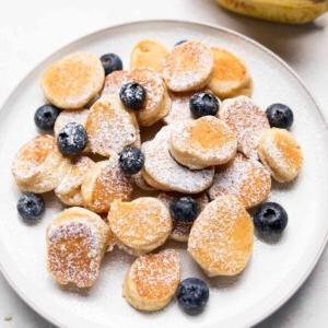 Mini Bocconcini di Pancake alla Banana 4 mini pancake di banana guarniti con zucchero a velo e mirtilli serviti su un piatto bianco