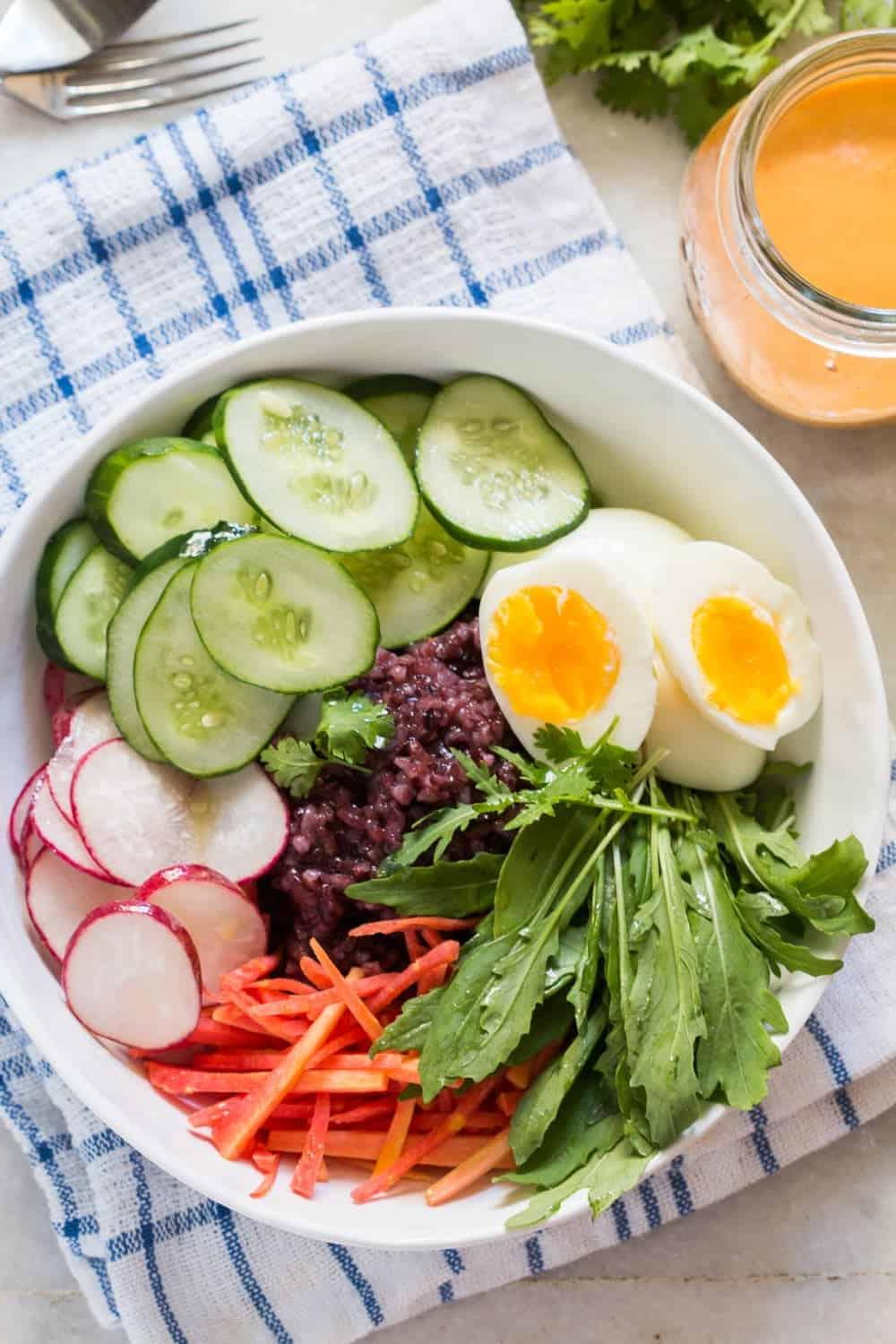 **Ciotola di Insalata di Riso Venere con Condimento Piccante alle Arachidi** 5 vista dall'alto degli ingredienti per l'insalata di riso nero in una ciotola bianca su una tovaglia blu e bianca con un barattolo di salsa sul lato