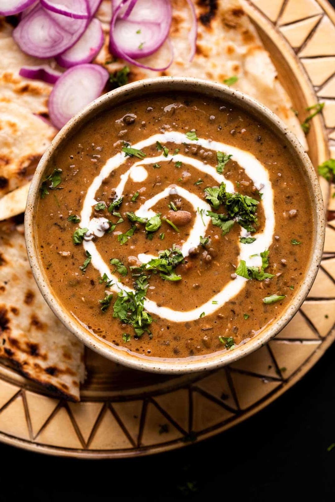 Dal Makhani – La Ricetta Definitiva per Fornello e Pentola a Pressione 6 Dal Makhani servito in una ciotola di ceramica marrone con panna versata sopra e naan e cipolle affettate a lato
