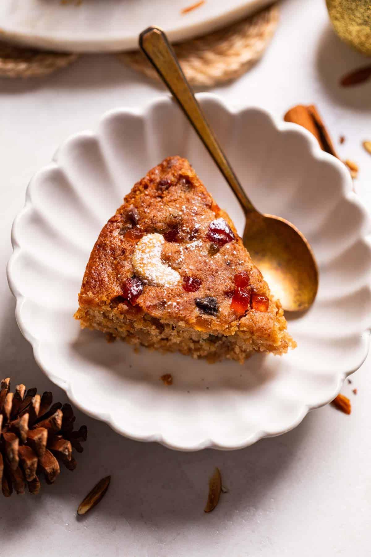Plumcake Senza Uova 6 Una fetta di torta di prugne senza uova su un piatto bianco, accompagnata da un cucchiaio, pronta per essere assaporata. Si possono vedere i ricchi frutti secchi all'interno della mollica umida.