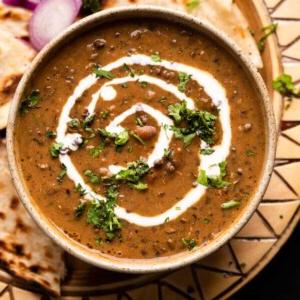 Dal Makhani – La Ricetta Definitiva per Fornello e Pentola a Pressione 7 Dal Makhani servito in una ciotola di ceramica marrone con panna versata sopra e naan e cipolle affettate a lato