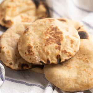 Ricetta del Pane Pita Integrale 8 Immagine di pani pita integrali appena sfornati, disposti su un panno da cucina a righe bianche, caldi e pronti per essere gustati immediatamente.