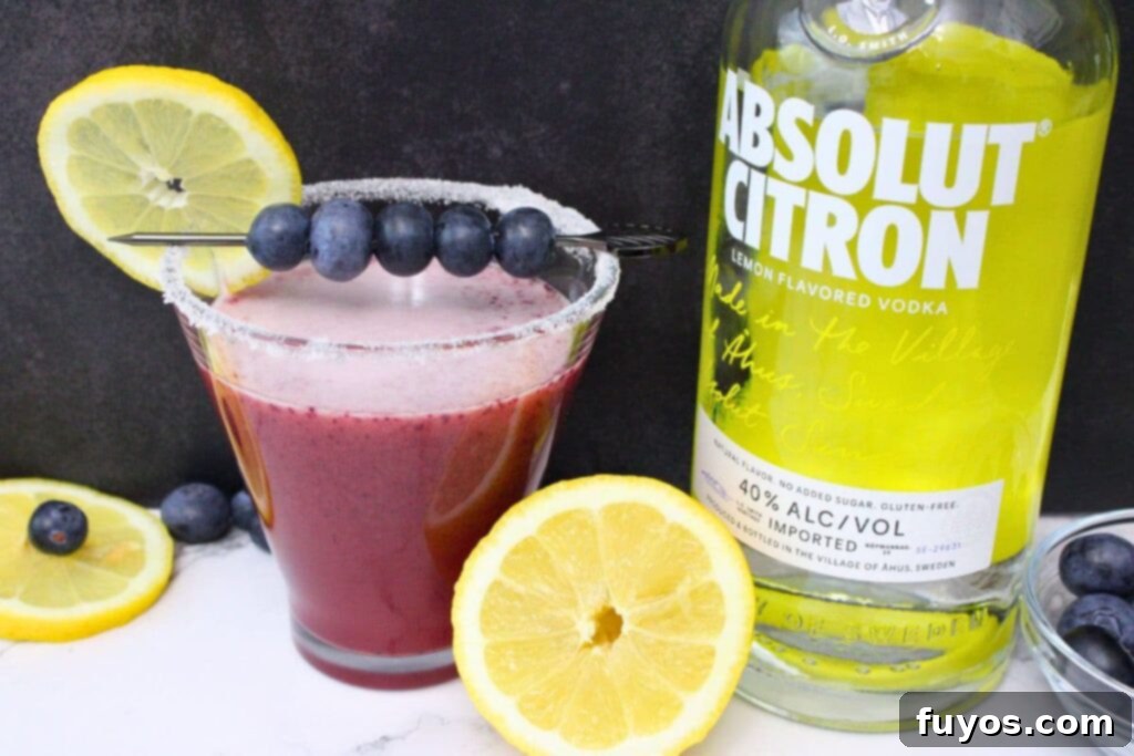 A glass of blueberry lemon drop martini beside a bottle of Absolut Citron vodka, highlighting a key ingredient.
