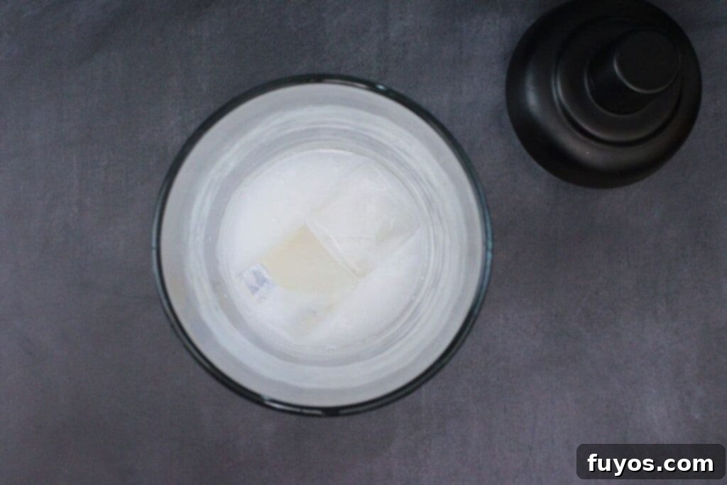 A close-up of a cocktail shaker being filled with coconut rum, vanilla vodka, and coconut cream, with ice cubes already inside, preparing for a Drunk Ghost Cocktail.