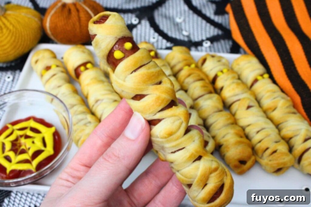 Air Fryer Mummy Dogs Your Easy Halloween Fun 9 holding up a mummy hot dog made in the air fryer with mustard eyes