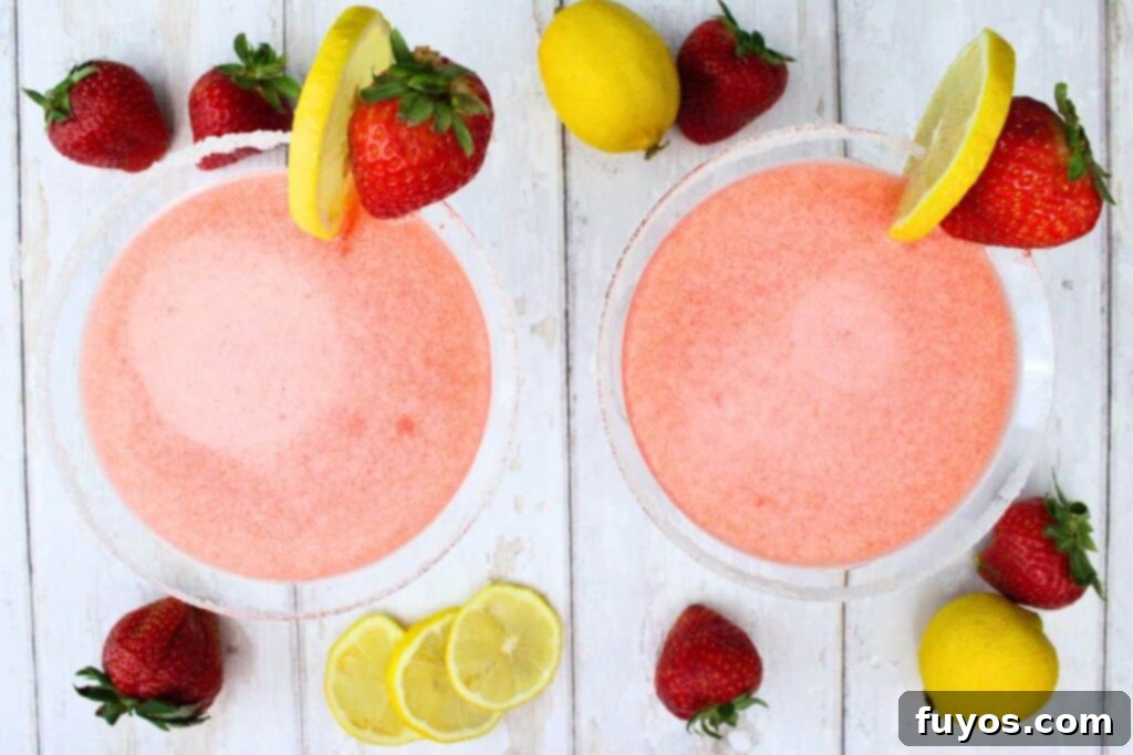 An overhead view of two completed strawberry lemon drop martinis, garnished with lemon and strawberry, on a white wooden table surrounded by fresh fruit.