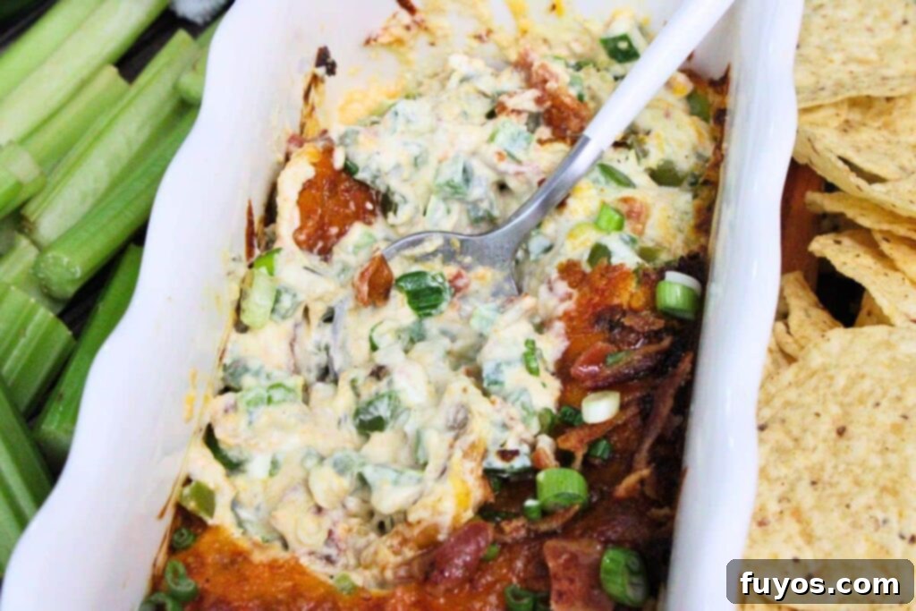 A dish of creamy jalapeno popper dip with a serving spoon, garnished with green onions, next to fresh celery sticks and tortilla chips.