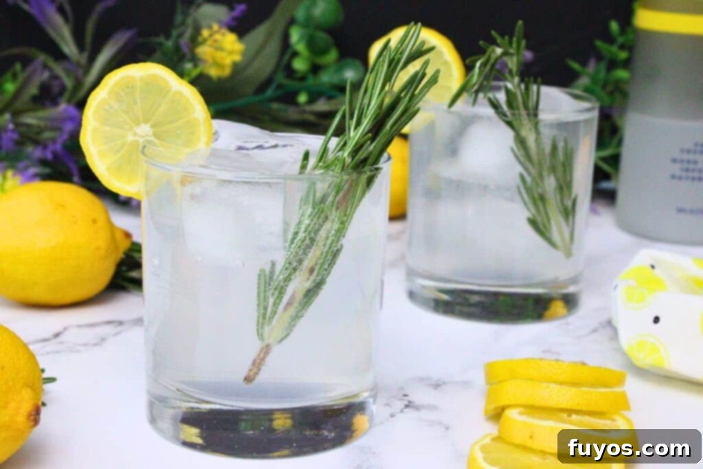 two sparkling lemonade cocktails, one in foreground with rosemary and lemon, surrounded by fresh lemons and white flowers