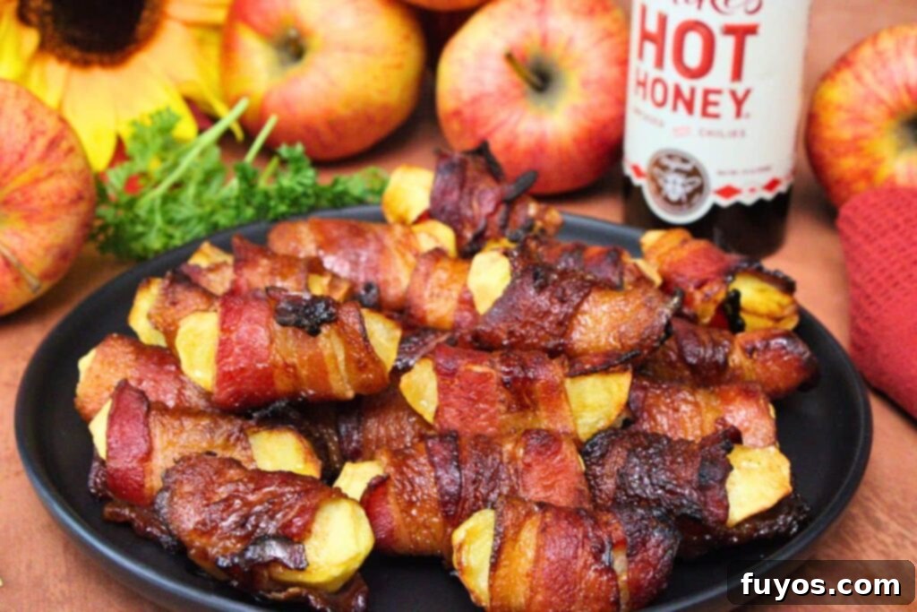 Sweet and Savory Air Fryer Bacon Apple Bites 9 bacon wrapped apples stacked on a black plate in front of fresh apples and hot honey bottle