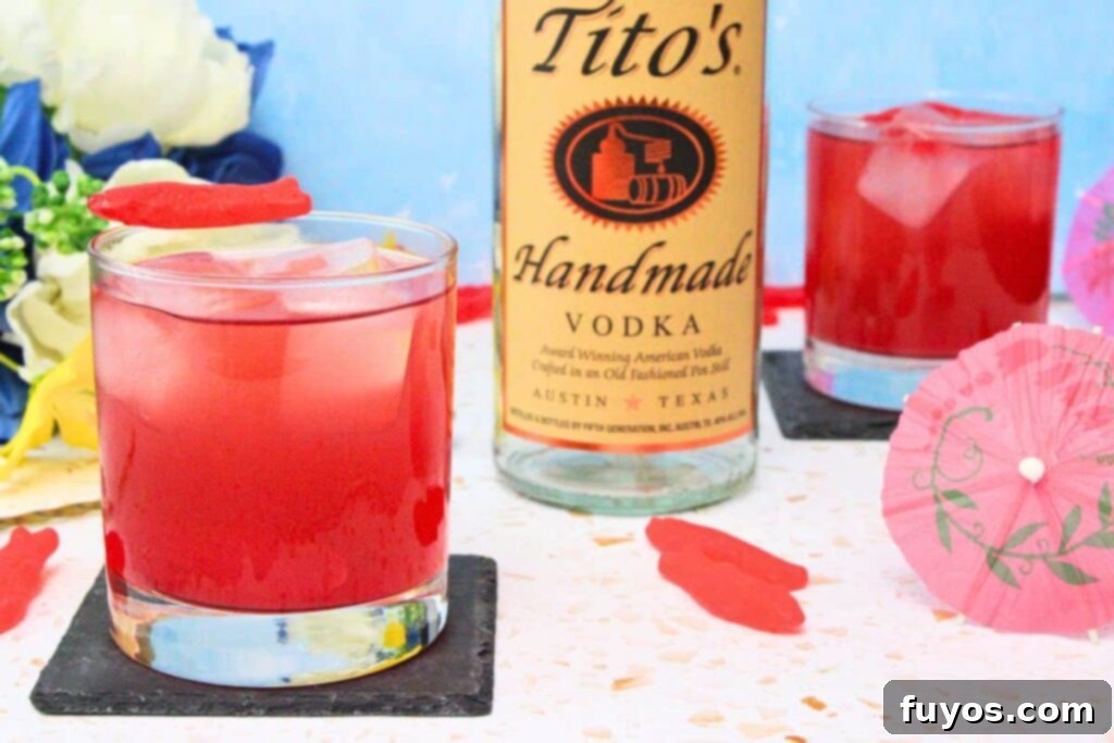 A Swedish Fish cocktail on ice, garnished with gummy candies, next to an infused vodka bottle on a bright, summer-themed background.