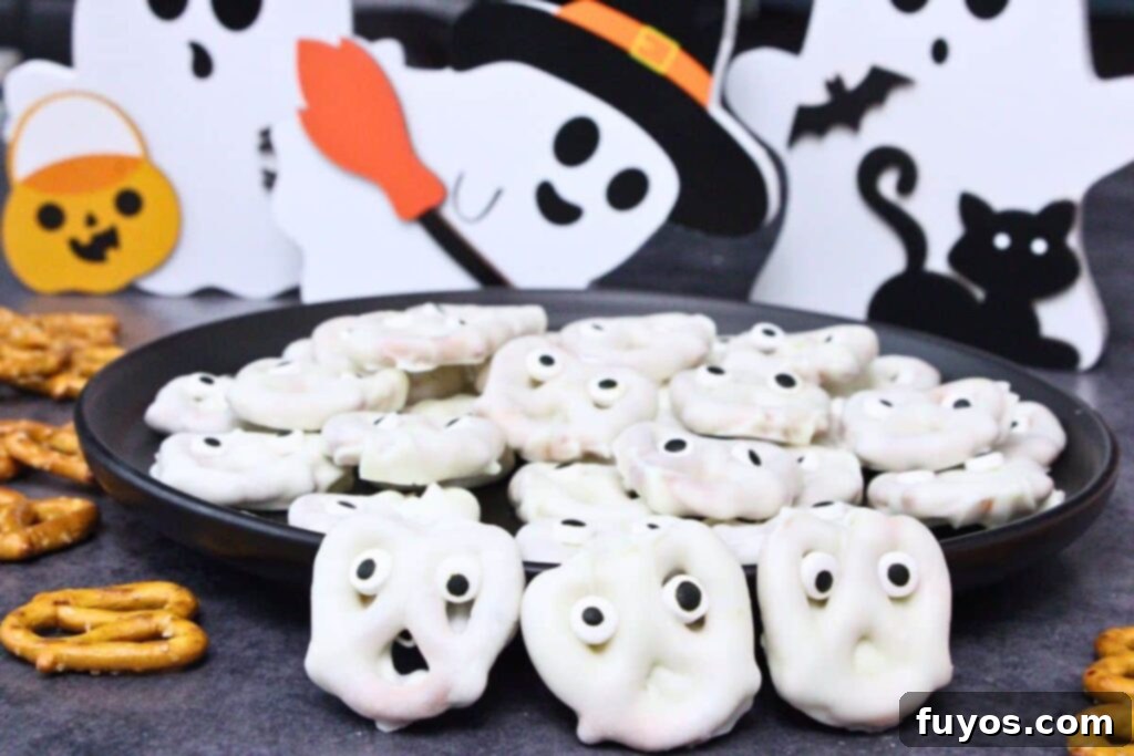 white chocolate covered ghost pretzels in front of a plate of more ghost pretzels surrounded by halloween decorations