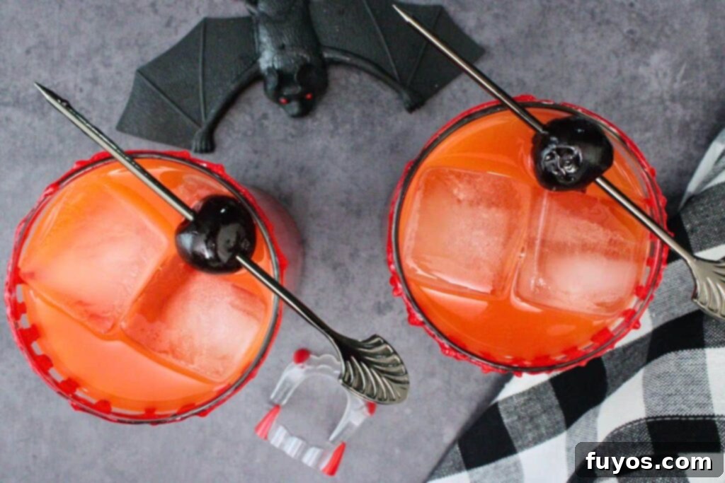 overhead view of two vampire tequila sunrise drinks with cherry skewer on a dark counter with bat and vampire fang decorations