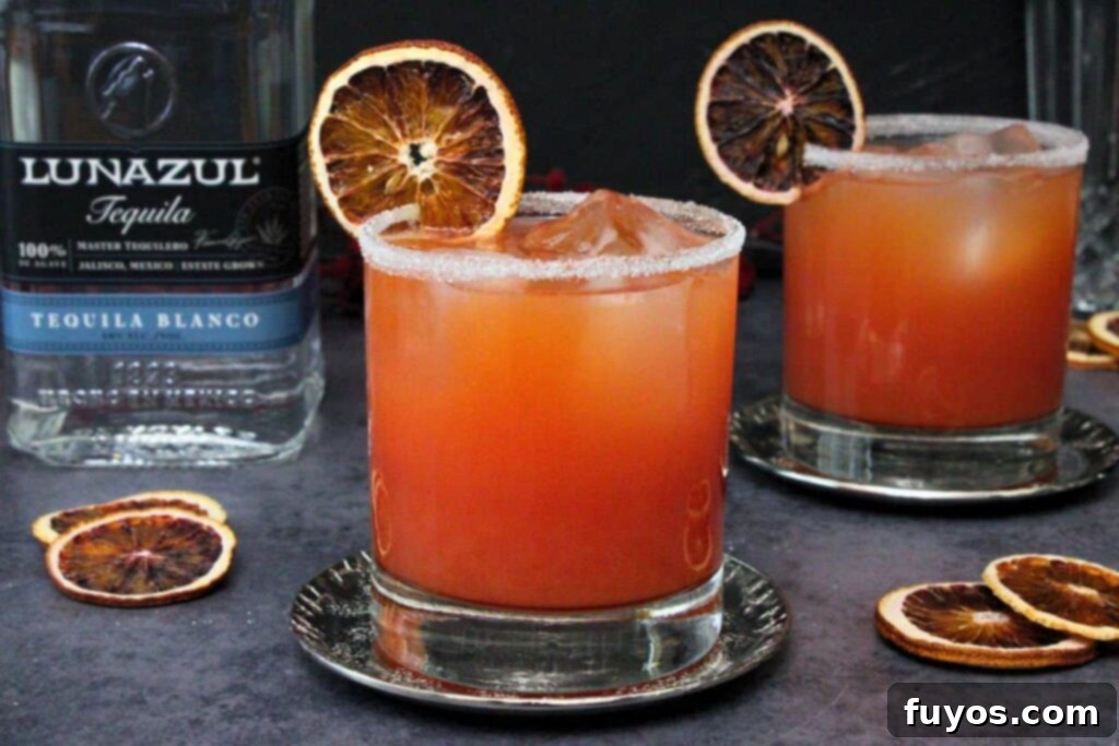 Blood Orange Margaritas The Perfect Spooky Cozy Sip 3 blood orange margarita with blood orange slice and sugar rim on a metal coaster with separate drink and tequila bottle in background