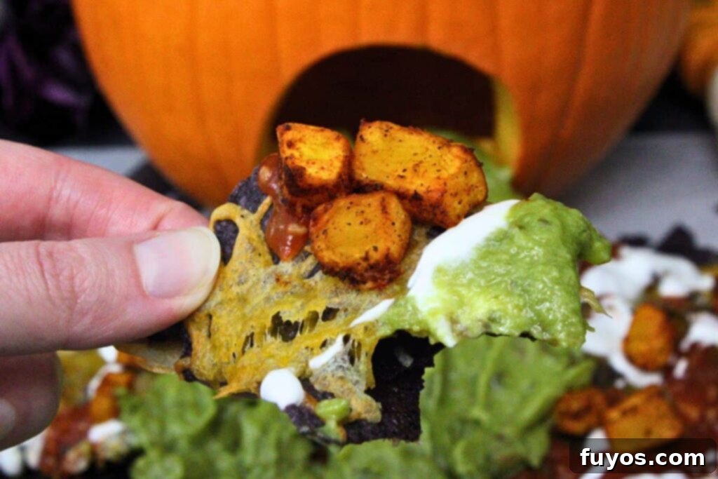 Air Fryer Halloween Monster Pumpkin Nachos 3 holding up a halloween nacho showing the blue corn tortilla chip with air fryer diced pumpkin, guacamole, salsa, cheese, and sour cream
