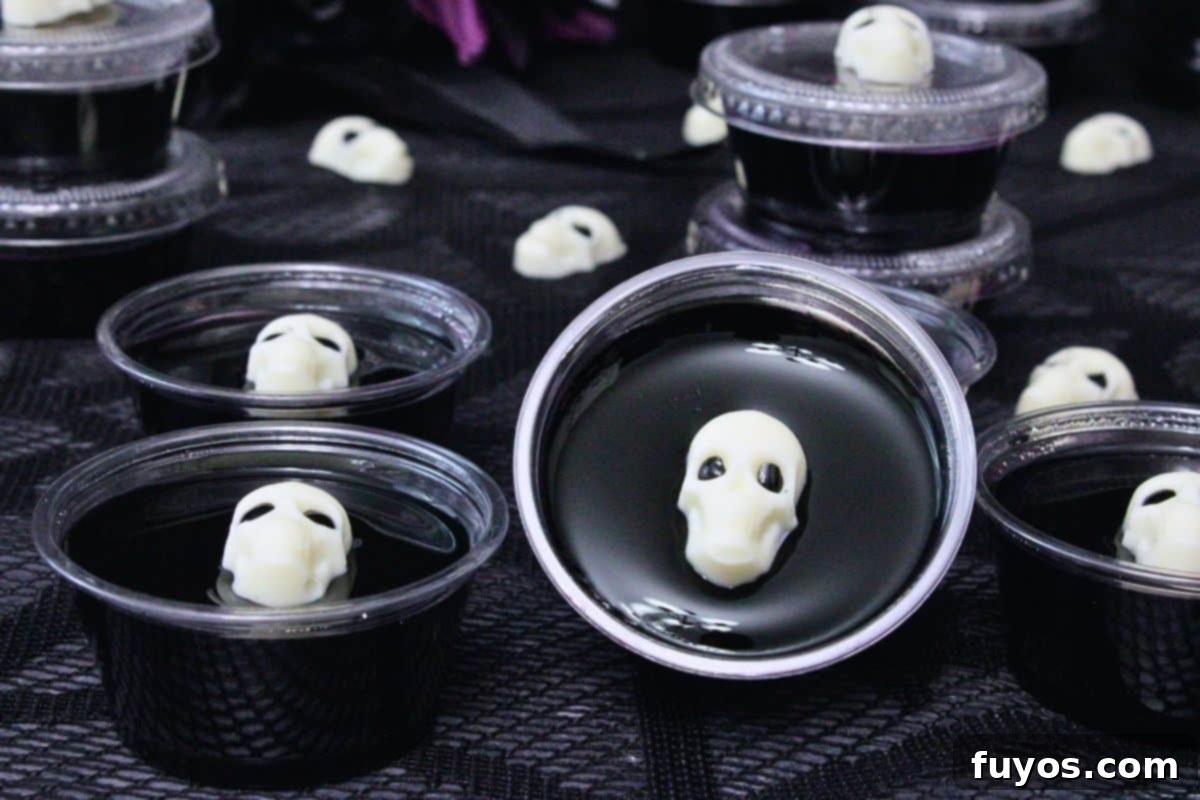 closeup of several black jello shots in plastic cups topped with a white chocolate skull on a black lace web cloth