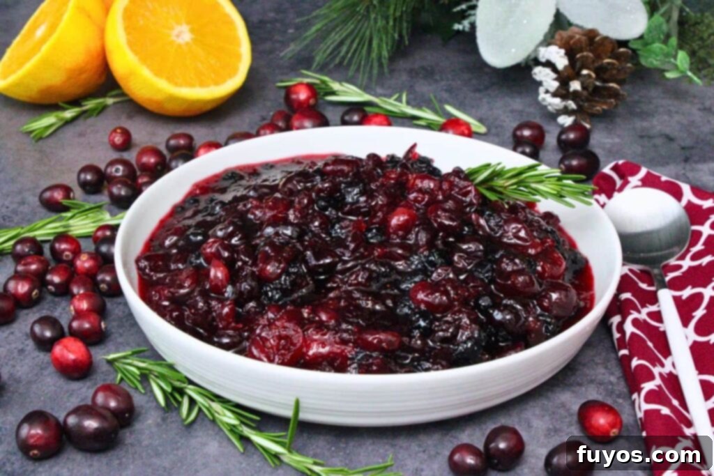 Bowl of glistening microwave cranberry sauce with a sprig of fresh rosemary, surrounded by whole cranberries, oranges, and more rosemary on a rustic surface