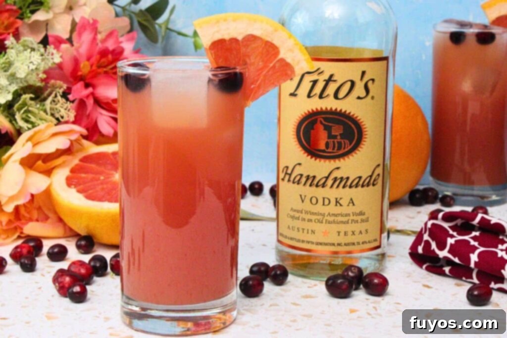 Sea Breeze cocktail in a tall glass with grapefruit wedge garnish next to vodka bottle amidst fresh fruit and flowers