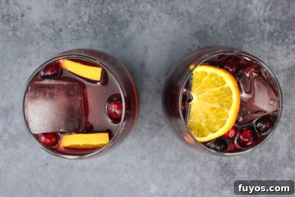A cranberry sangria drink being filled with ice cubes, fresh cranberries, and orange slices, illustrating the second step of preparation.