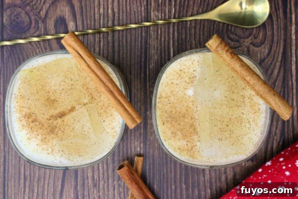Overhead view of two amaretto eggnog cocktails garnished with nutmeg and cinnamon sticks on a wooden counter