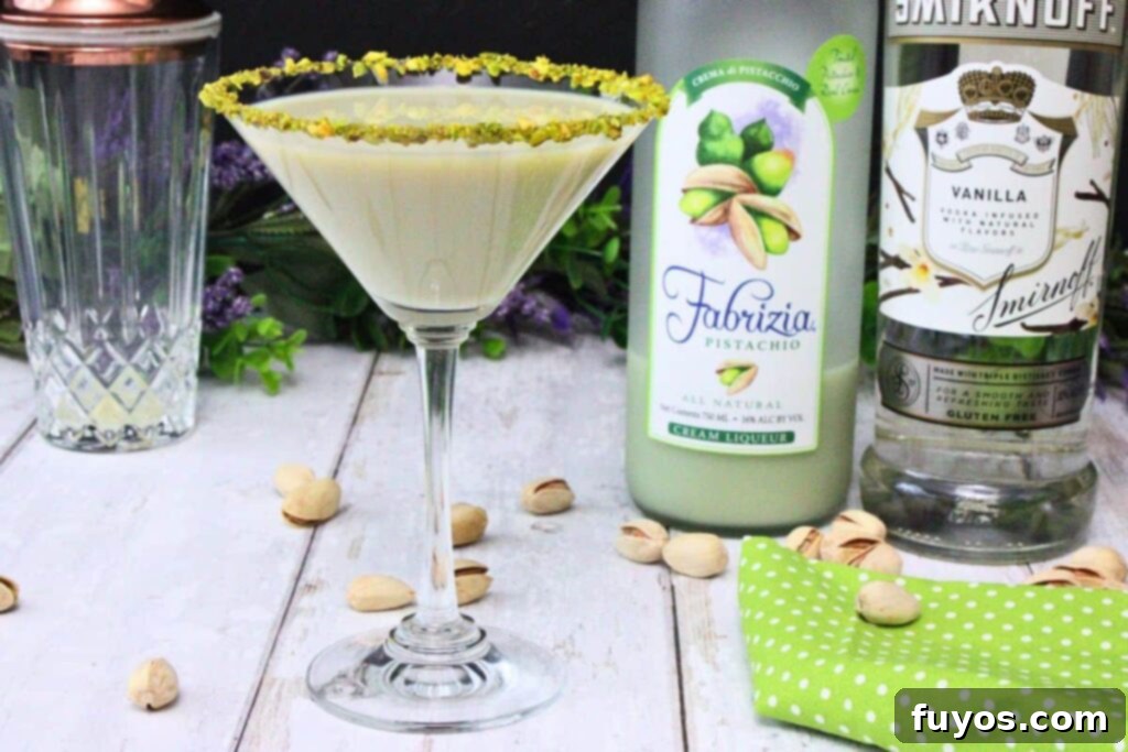 pistachio martini with rimmed glass in front of liquor bottles on a white wooden counter sprinkled with pistachios